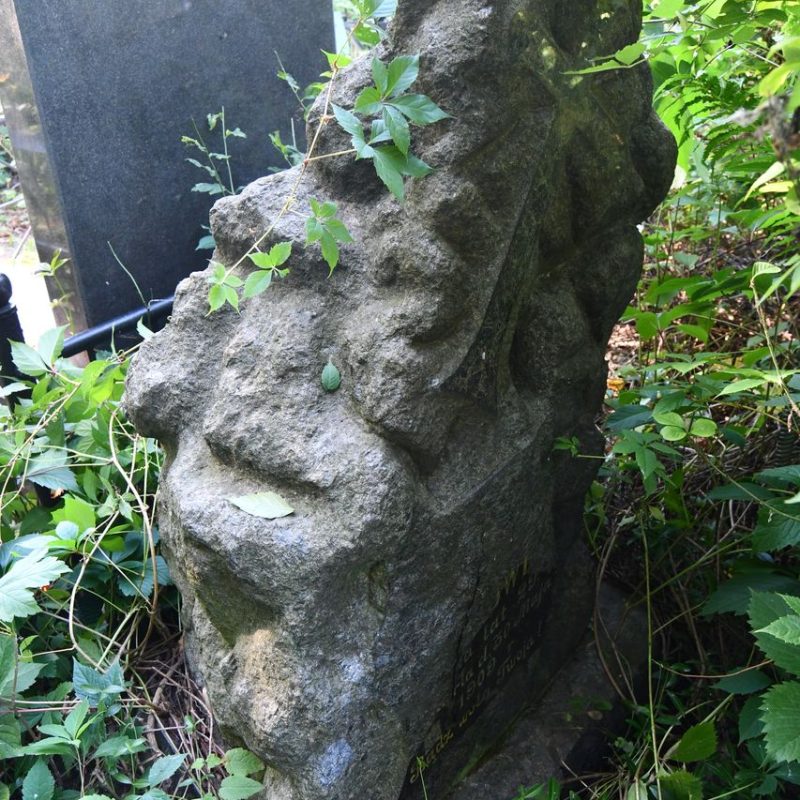 Pierre tombale avec un socle stylisé comme un tas de pierres, avec une plaque en relief portant une inscription et une croix avec un bras inférieur élargi et des extrémités semi-circulaires.