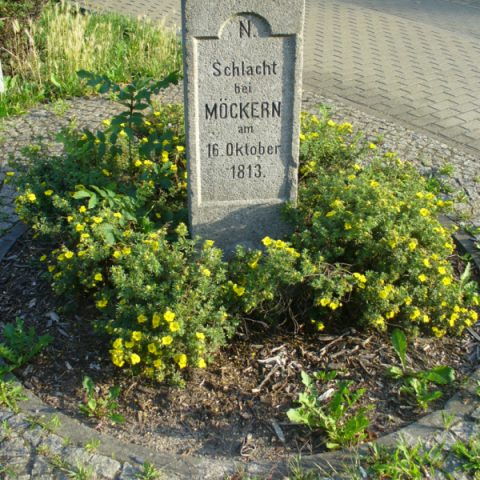 Kamienny pomnik w Lipsku upamiętniający bitwę pod Möckern 16 października 1813 roku, otoczony żółtymi kwiatami. Napis: 'Schlacht bei Möckern am 16. Oktober 1813'.