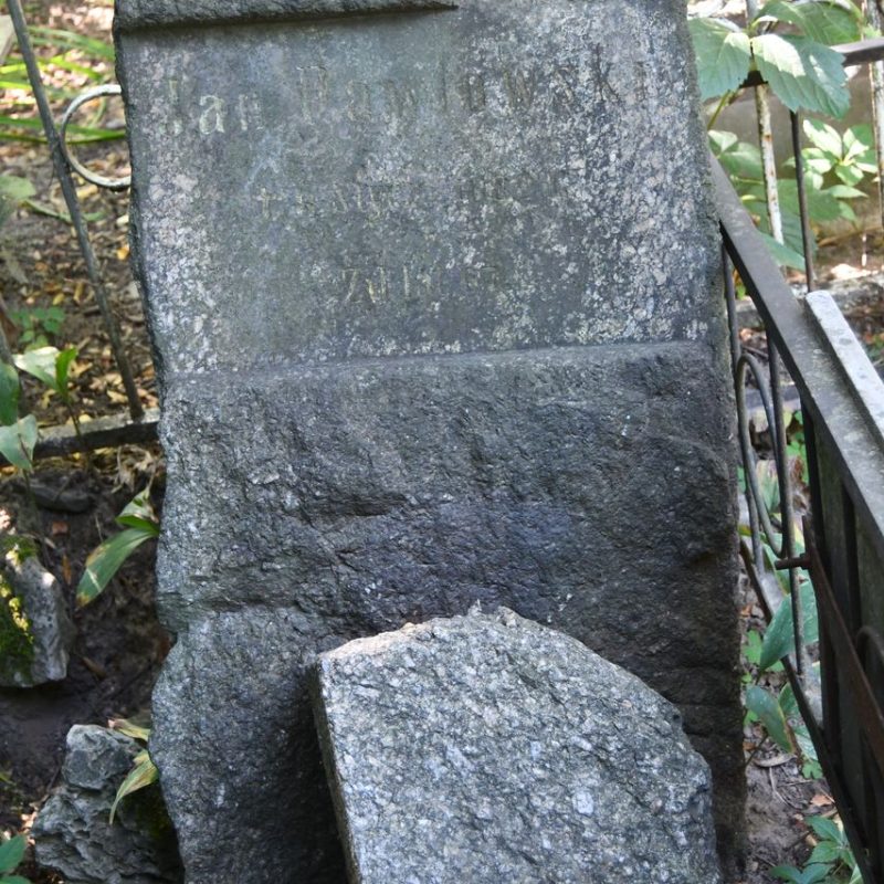 Stone pedestal with an inscription for Jan Pawłowski, missing its cross. The pedestal is a rough block with engraved text on the front. Surrounded by vegetation and metal fencing.