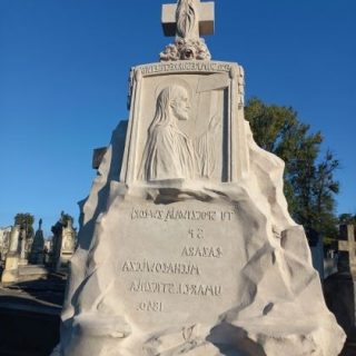 White stone tombstone with a relief of a saint and inscriptions in Cyrillic, topped with a cross, in a cemetery under a clear blue sky.