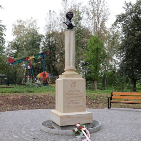 Pomnik Adama Mickiewicza w Borysławiu, Ukraina, z brązowym popiersiem na kanelowanej kolumnie i marmurową tablicą. Pomnik otoczony drzewami, w tle widoczna kolorowa konstrukcja placu zabaw.
