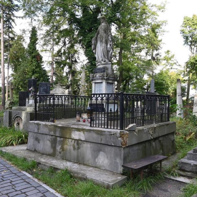 Nagrobek na Cmentarzu Łyczakowskim we Lwowie, otoczony czarnym metalowym ogrodzeniem. Na szczycie nagrobka stoi figura kobiety, w tle drzewa i inne groby.