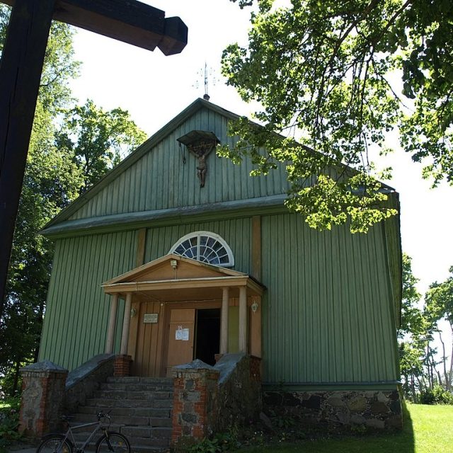 Église en bois de Saint-Casimir à Powiewiórka, Lituanie, avec une façade verte, des marches à l'avant et une croix partiellement visible à gauche. Des arbres entourent l'église.