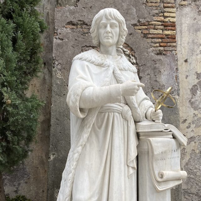 A marble sculpture of Nicolaus Copernicus’s head, featuring facial details and flowing hair. Greenery and flowers in the background.