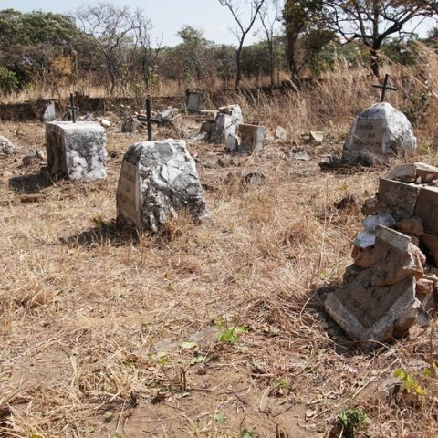 Tombes dans la section polonaise du cimetière à Mbala, Zambie. La zone est envahie par de l'herbe sèche, et plusieurs pierres tombales avec des croix sont visibles. Des arbres et un mur de briques entourent le cimetière.