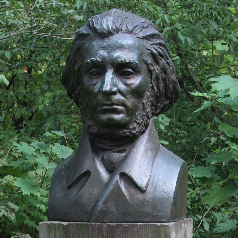 Buste en bronze d'Adam Mickiewicz dans un parc, entouré de feuillage vert dense. La sculpture montre Mickiewicz avec une expression sérieuse, des cheveux détaillés et un manteau à col haut.