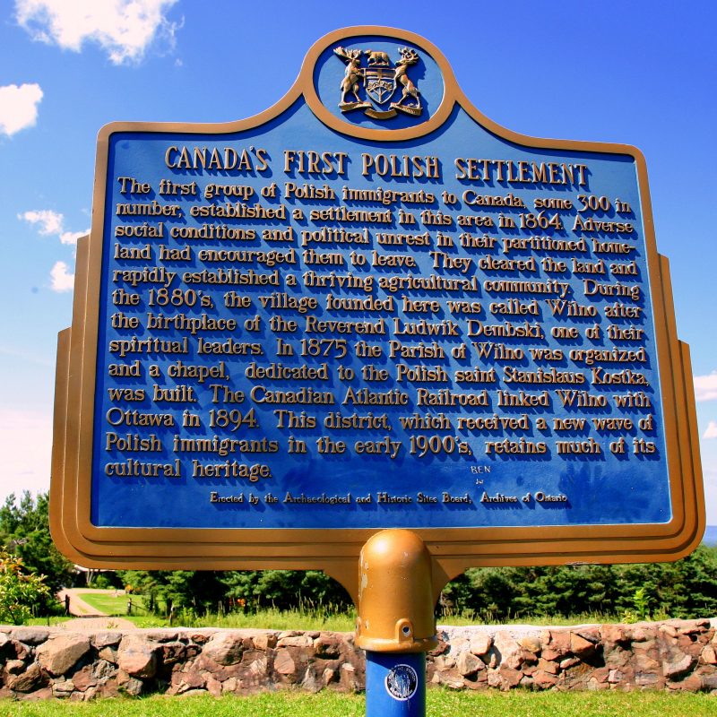 A blue historical plaque entitled ‘The First Polish Settlement in Canada’, featuring text describing the establishment of the Polish community in Wilno, Ontario, in the 1860s. The plaque is set against a backdrop of trees and a clear sky.