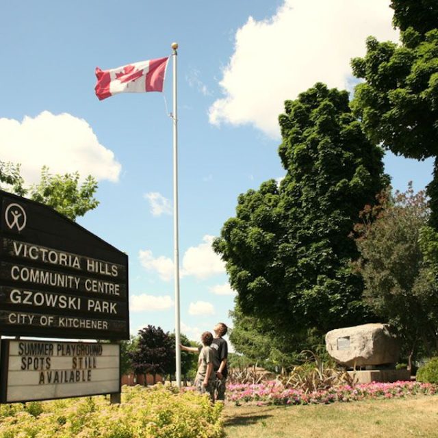 Park Gzowskiego w Kitchener z kanadyjską flagą na maszcie, tablicą z napisem 'Victoria Hills Community Centre Gzowski Park City of Kitchener' oraz dużym kamiennym pomnikiem otoczonym drzewami i kwiatami.