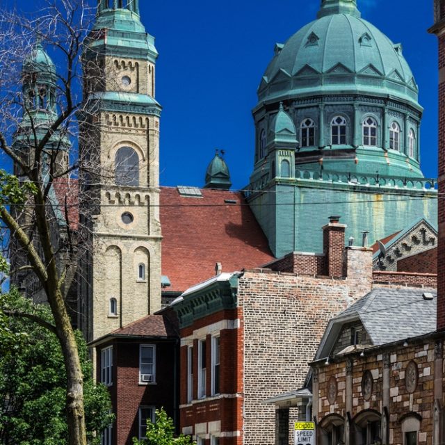 Kościół Matki Bożej Nieustającej Pomocy w Chicago z neoromańską fasadą, smukłymi wieżami i zielonymi kopułami. Kościół otoczony jest budynkami mieszkalnymi i ulicą z drzewami.