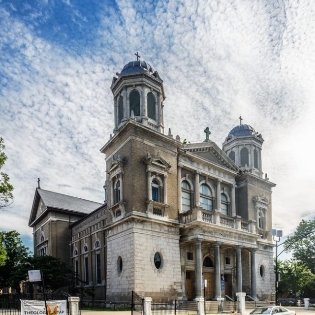 Kościół św. Jadwigi Śląskiej w Chicago z charakterystyczną fasadą z dwoma wieżami i portykiem. Budynek zbudowany z żółtobrązowej cegły i detali z piaskowca z Bedford, pod częściowo zachmurzonym niebem.
