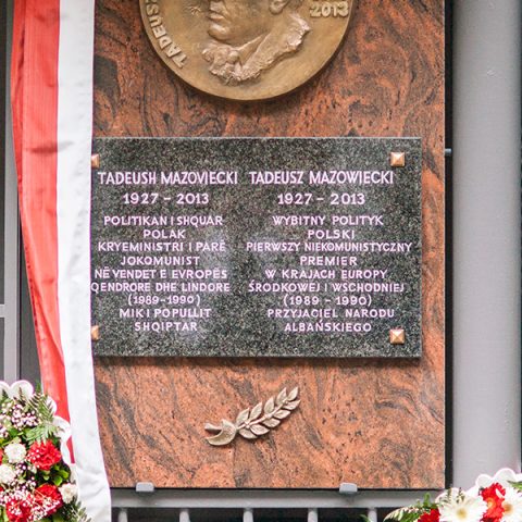 Bronze relief plaque commemorating Tadeusz Mazowiecki in Tirana, featuring his portrait and bilingual inscription in Polish and Albanian. Surrounded by flowers and a red and white ribbon.