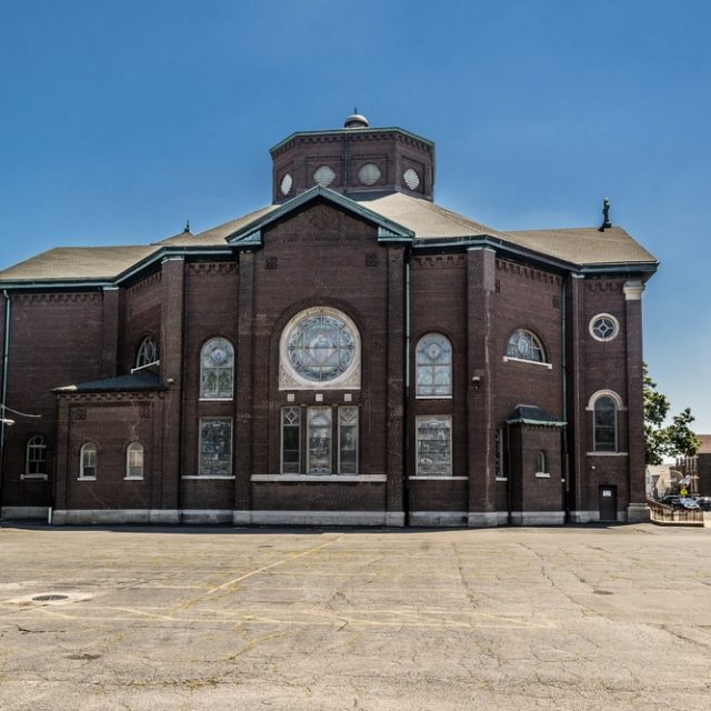 Église en brique de Sainte-Barbara à Chicago avec un plan central et un toit octogonal, dotée de grandes fenêtres en vitrail et d'une façade simple. La zone environnante est pavée avec des arbres sur les côtés.