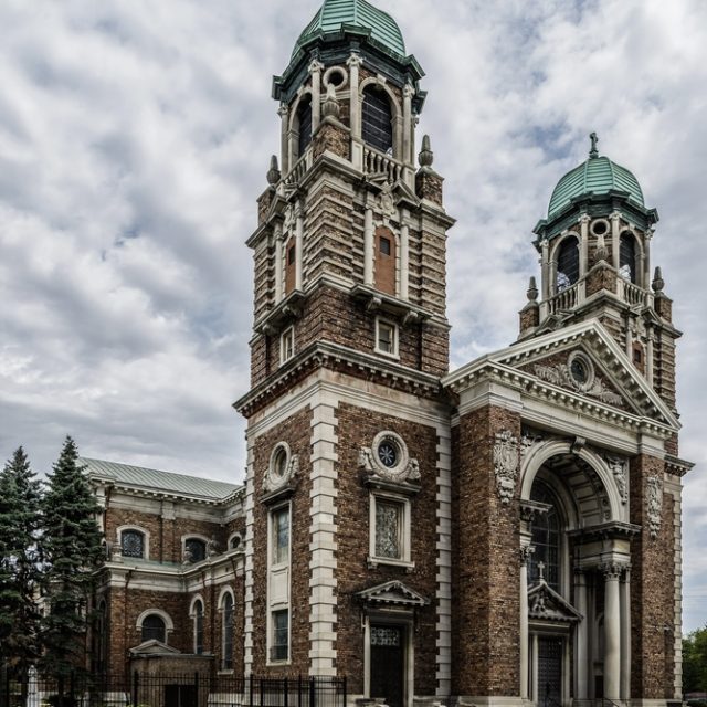 St. Francis of Assisi Church in Detroit with two square towers and a large arch on the facade. The building is made of brick with stone details. Trees and a fence are visible nearby.