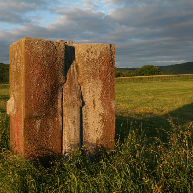 Adolf Ryszka's sculpture 'Boundary Stone' made of red sandstone, standing on a grassy hill in Sankt Wendel, with a split in the center, surrounded by fields and distant hills.