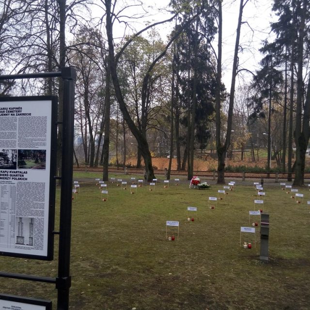 Widok na kwaterę żołnierzy polskich na cmentarzu na Zakrecie z tablicą w kilku językach oraz rzędami małych tabliczek i świec na trawie.