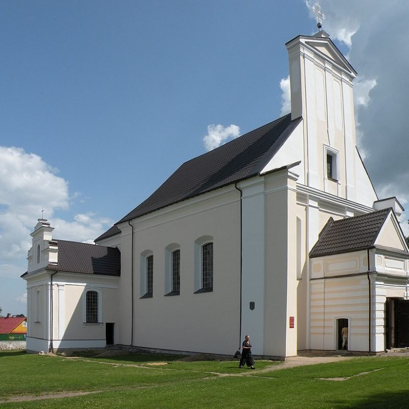 L'église Saint-Nicolas à Gieranony, Biélorussie, présente une façade blanche avec un toit sombre, des éléments baroques et un petit porche d'entrée. Le bâtiment est entouré de pelouse verte et d'un ciel dégagé.