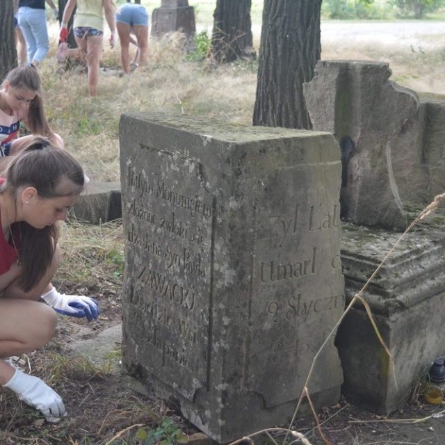 Młodzi wolontariusze sprzątają wokół zniszczonego nagrobka z inskrypcjami na polskim cmentarzu na Ukrainie. Obok kamienia stoją znicze.