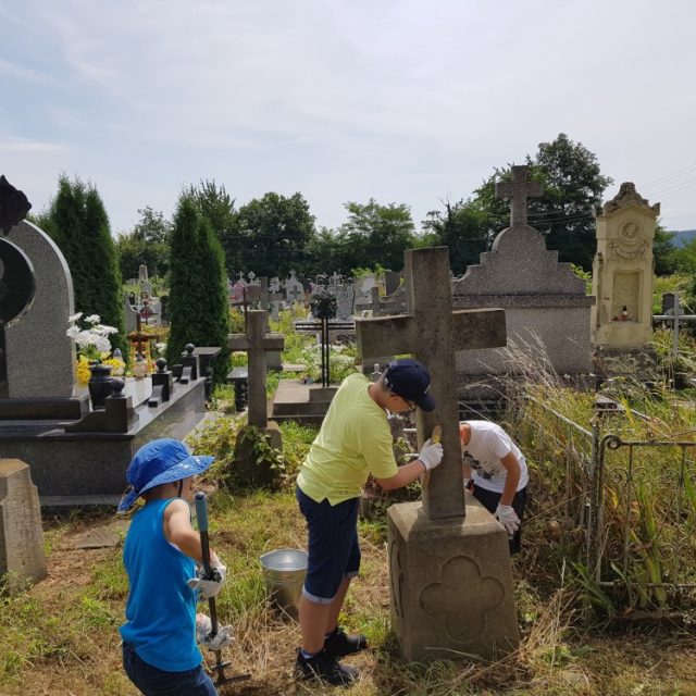 Volunteers cleaning gravestones at an Armenian cemetery in Ukraine. Two people work on a stone cross, while another tends to the area. Various gravestones and greenery are visible in the background.