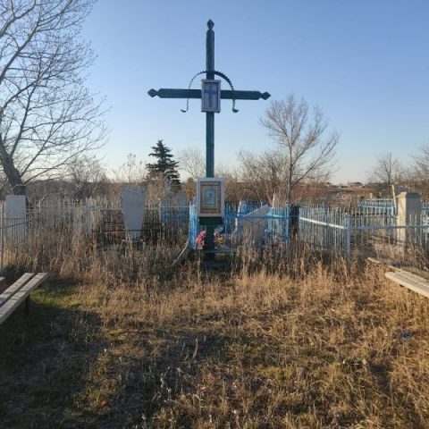 Un cimetière au Kazakhstan avec une grande croix en bois entourée d'herbes hautes et de plusieurs pierres tombales. La zone est entourée d'une clôture métallique, et des arbres sont visibles en arrière-plan.