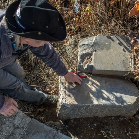 Людина в капелюсі чистить зношений надгробок, оточений сухою рослинністю на кладовищі Кукія в Тбілісі.