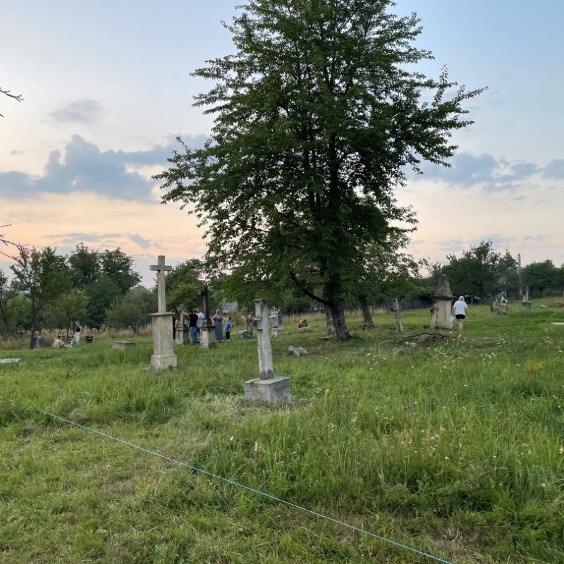 Vue du cimetière polonais à Cacica, Roumanie, avec plusieurs anciennes pierres tombales et croix dispersées sur une zone herbeuse. Un groupe de personnes est rassemblé près des tombes sous un arbre au coucher du soleil.