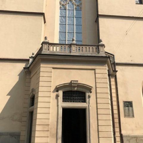 The western porch of the Latin Cathedral in Lviv, featuring a tall, arched window with decorative tracery above the entrance. The facade is light-colored with simple architectural details.