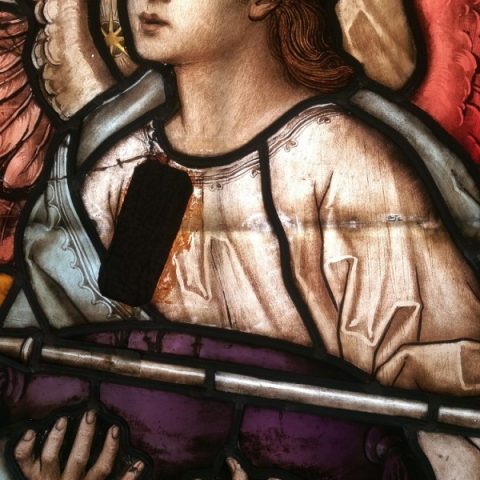 Detail of the stained glass 'Our Lady Queen of Angels' in Lviv Cathedral, showing an angel with red wings holding a purple scroll. The angel's face is serene, and there is a missing glass piece on the chest.