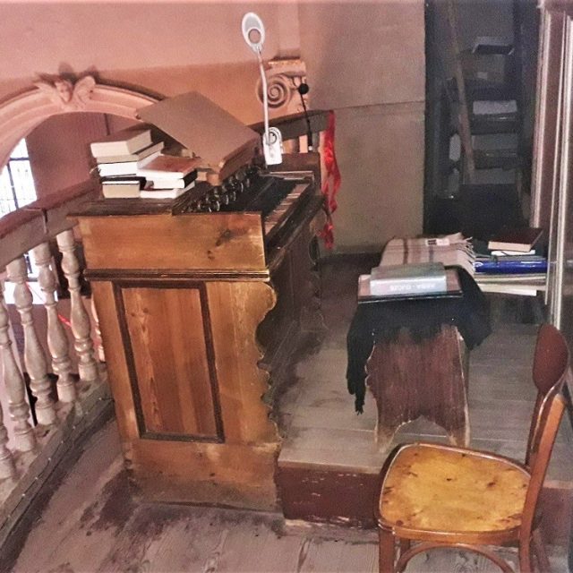 Widok na konsolę organową i chór muzyczny w kościele w Krzemieńcu. Drewniana konsola otoczona jest balustradą, obok znajdują się książki i krzesło.