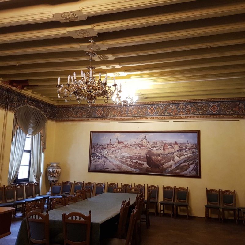 Intérieur de la salle représentative de la Maison Royale à Lviv. La pièce présente un plafond en bois avec des éléments décoratifs, une frise florale, un grand lustre et une longue table entourée de chaises. Deux fenêtres avec rideaux drapés sont visibles.