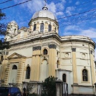 Kościół Świętych Piotra i Pawła w Tbilisi, Gruzja, z charakterystyczną kopułą i żółtą fasadą, pod niebieskim niebem z chmurami. Na pierwszym planie widoczne drzewa i ogrodzenie.