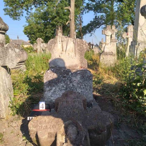 Old gravestones at Zbarazh cemetery surrounded by overgrown grass and trees, with a numbered marker indicating restoration work.