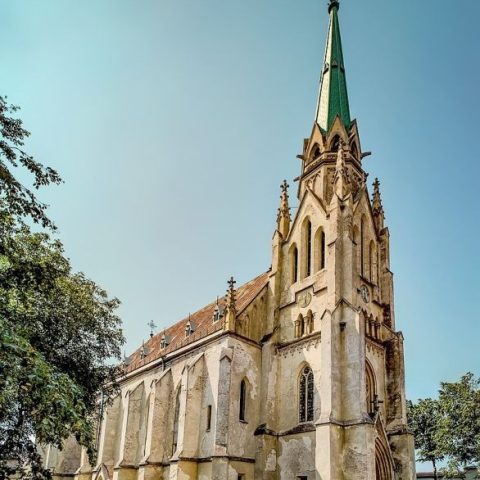 Église néogothique du Sacré-Cœur de Jésus à Tchernivtsi, Ukraine, avec une haute flèche et des arcs pointus, entourée d'arbres et d'un ciel dégagé.