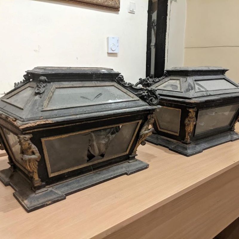 Deux reliquaires baroques avec des éléments décoratifs sur une table en bois. Les reliquaires ont des panneaux de verre et des détails métalliques ornés, y compris des figures aux coins.