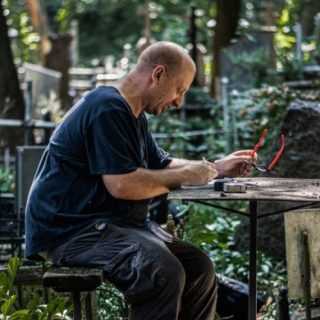 Un homme assis à une table dans un cimetière, tenant des lunettes rouges, entouré de pierres tombales et de verdure.