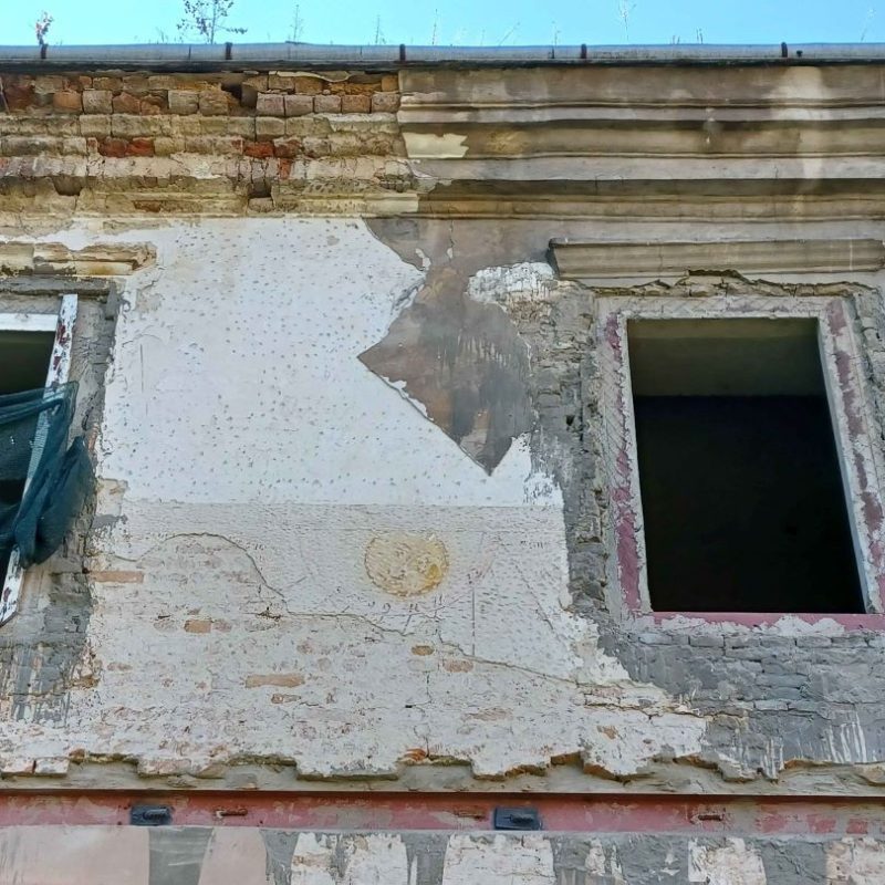 Zewnętrzna ściana zamku w Żółkwi z częściowo zachowaną polichromią przedstawiającą motyw zegara słonecznego. Ściana jest uszkodzona, widoczne cegły i dwa otwory okienne, jeden zasłonięty siatką.