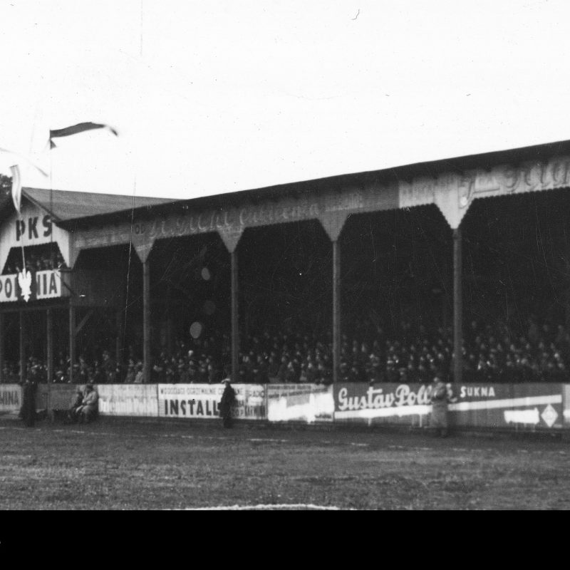 Black and white photo of a crowded grandstand at PKS Polonia Karwina stadium. Banners and advertisements are visible on the structure. A few people stand on the field in front of the grandstand.