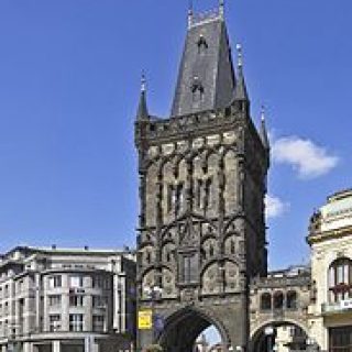 Brama Prochowa w Pradze, gotycka budowla z misternymi rzeźbieniami w kamieniu, wznosi się na tle błękitnego nieba. Obok przechodzą ludzie, podkreślając jej historyczne znaczenie.