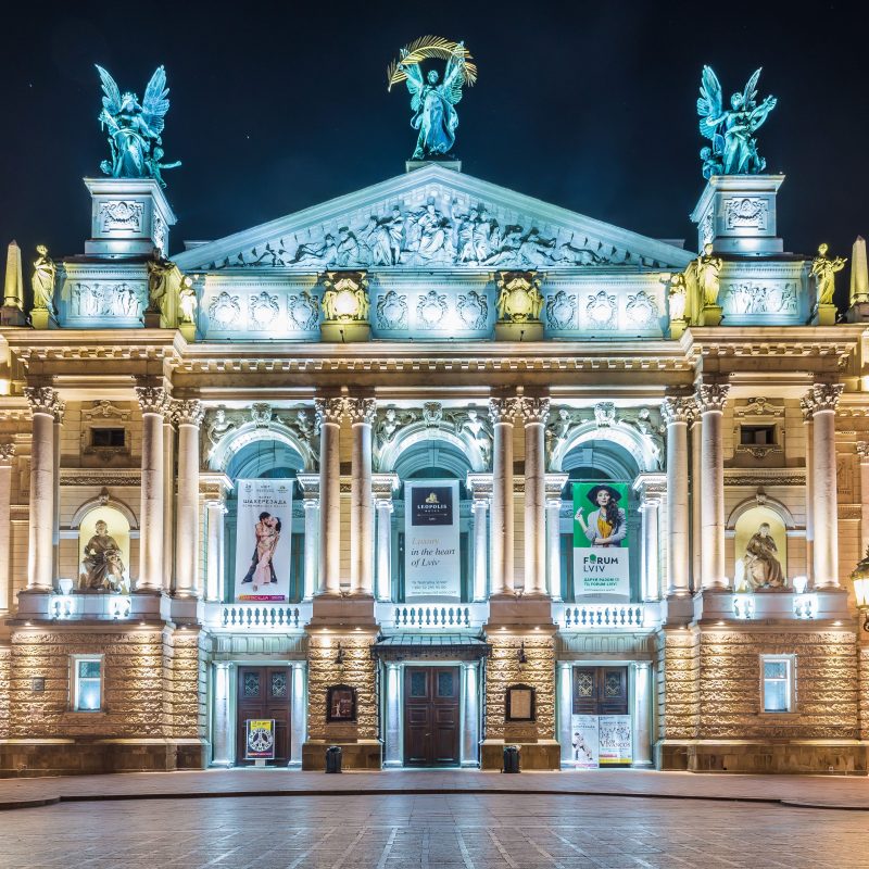 Vue nocturne de l'Opéra de Lviv, conçu par Zygmunt Gorgolewski. La façade illuminée présente des colonnes, des statues et des bannières. Deux sculptures d'anges sont visibles sur le toit.