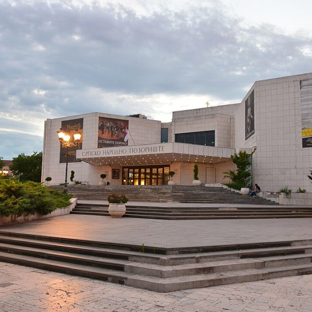 The National Theatre of Serbia building in Novi Sad, designed by Wiktor Jackiewicz, features a modern facade with large windows and banners. Steps lead to the entrance, surrounded by greenery and street lamps.