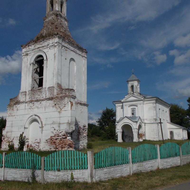The Catholic Church of Saints Peter and Paul in Dziewiątkowicze Nowe, Belarus, with a baroque bell tower and a white facade, surrounded by a green fence.