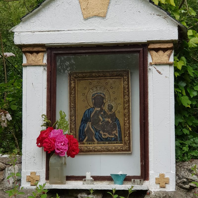 Mała kapliczka z ikoną Matki Boskiej z Dzieciątkiem, otoczona zielenią. Nad ikoną znajduje się symbol korony. Poniżej kwiaty w słoiku i małe świece.