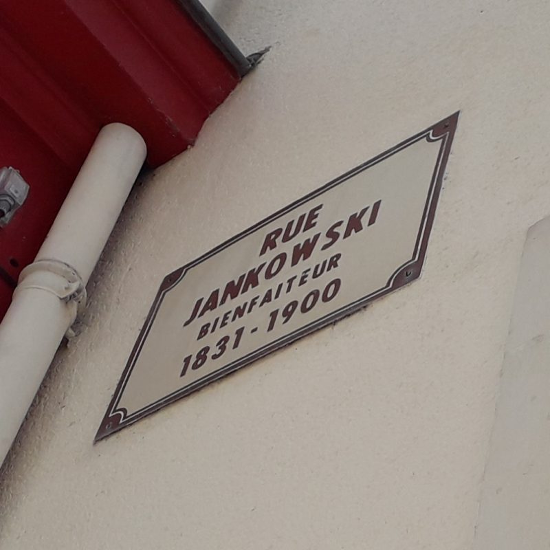 Tabliczka uliczna Rue Jankowski w Le Mans, upamiętniająca dobroczyńcę Filipa Jankowskiego, 1831-1900, zamontowana na ścianie obok czerwonej futryny drzwi.
