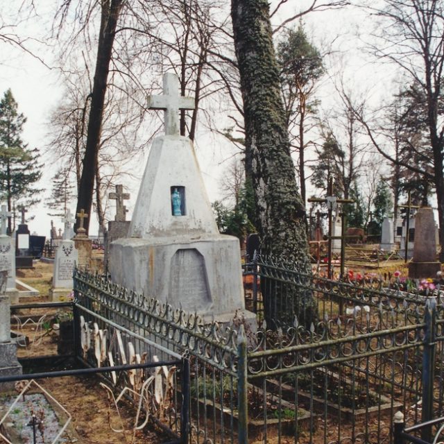 Grób otoczony ozdobnym metalowym ogrodzeniem na cmentarzu, z wysokim, białym nagrobkiem z krzyżem i małym obrazkiem. W tle widoczne są nagie drzewa.