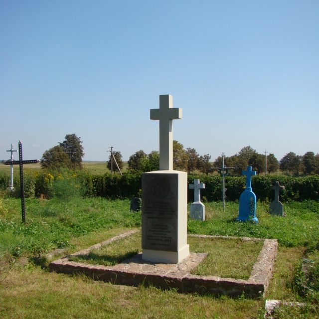 A concrete cross on a simple pedestal marks the grave of 18 Polish soldiers who died in September 1939, located in a grassy area with other graves in the background.