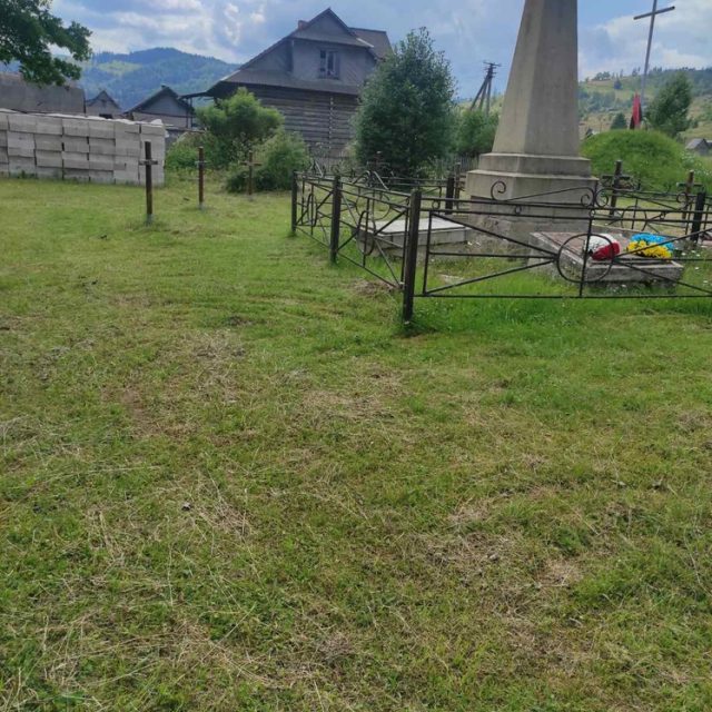 Пам'ятник II Бригаді Польських Легіонів у Рафайловій. Кам'яний обеліск з хрестом на вершині, оточений металевою огорожею. На задньому плані дерев'яний будинок і пагорби.