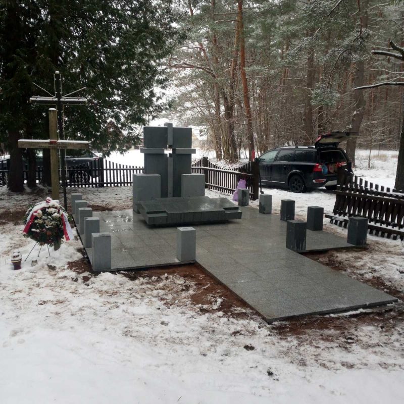 Un cimetière enneigé avec une croix en granit et une plaque commémorant les soldats polonais de la guerre polono-soviétique. Entouré d'arbres et d'une clôture en bois, avec une voiture garée à proximité.