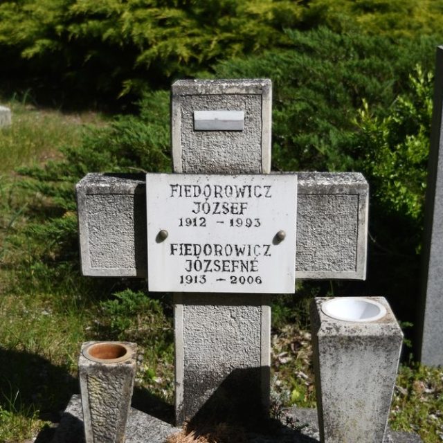 Tombe marquée par une croix avec une plaque portant les noms Józef et Józefa Fiedorowicz, dates 1912-1993 et 1913-2006. Un petit drapeau polonais est peint sur le bras supérieur. Deux vases cubistes encadrent la croix.