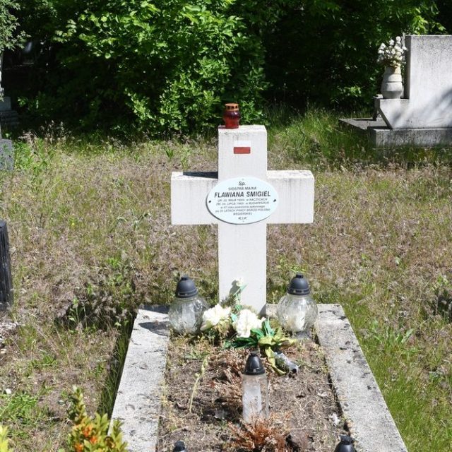 Grób oznaczony białym krzyżem z owalną tabliczką z nazwiskiem Marii Flawiany Smigiel. Na górnym ramieniu krzyża namalowana biało-czerwona flaga. U podstawy kwiaty i znicze.