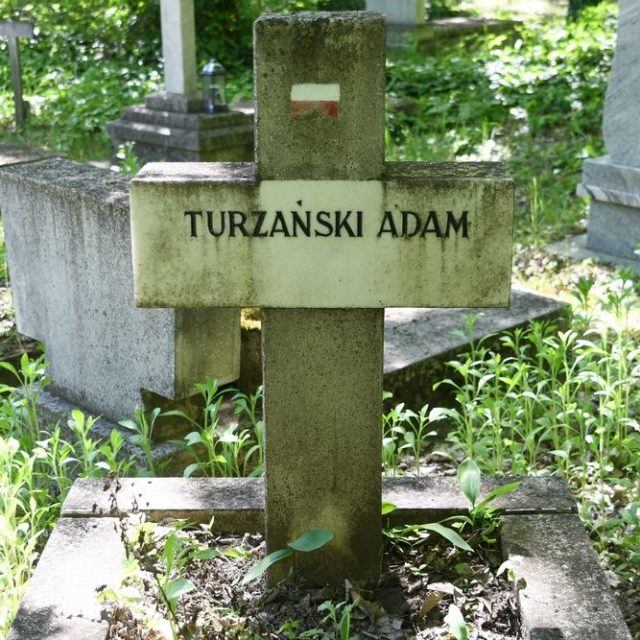 Могила, позначена хрестом з ім'ям Адам Туржанський. На горизонтальній балці табличка з написом, заповненим чорною фарбою. На верхньому плечі намальований біло-червоний прапор.