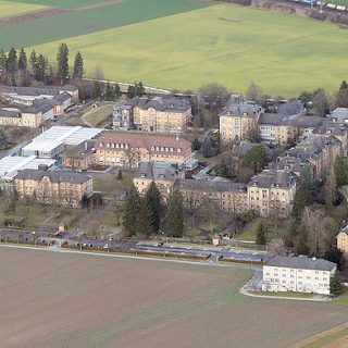 Vue aérienne de la clinique psychiatrique de Münsingen, montrant un complexe de bâtiments en brique entouré de champs et d'arbres. L'aménagement comprend des structures compactes et des pavillons séparés.